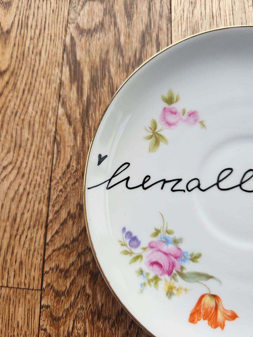 Weißer Vintage-Teller mit floralen Mustern und dem handgeschriebenen Wort „herzlich“.